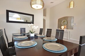 Un coin repas avec une table rectangulaire dressée pour six personnes, entourée de chaises. Un miroir est accroché au mur au-dessus de la table, et une petite plante est posée sur la table.