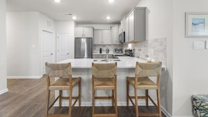 A modern kitchen with white cabinetry, a central island with bar stools, stainless steel appliances, and recessed ceiling lighting. - Belle Beach Retreat at Millville by the Sea (Millville)
