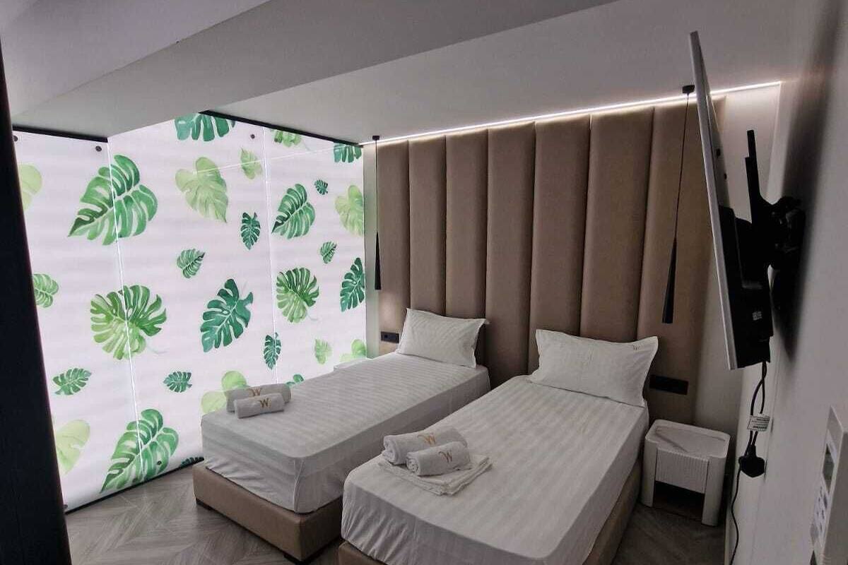 Habitació d'hotel moderna amb dos llits, una finestra gran amb un disseny de fulles i un paviment de fusta en relleu.