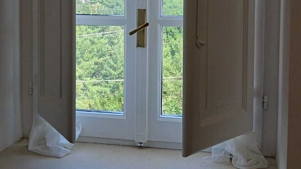 Quarto com uma grande janela mostrando duas portas brancas abertas, vista para colinas verdes e um radiador embaixo.