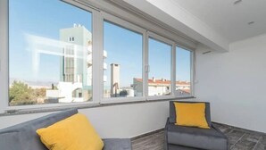 Un salón moderno con un sofá y un sillón grises, ambos con cojines amarillos, ventanas amplias que ofrecen vistas de edificios y un cielo despejado.