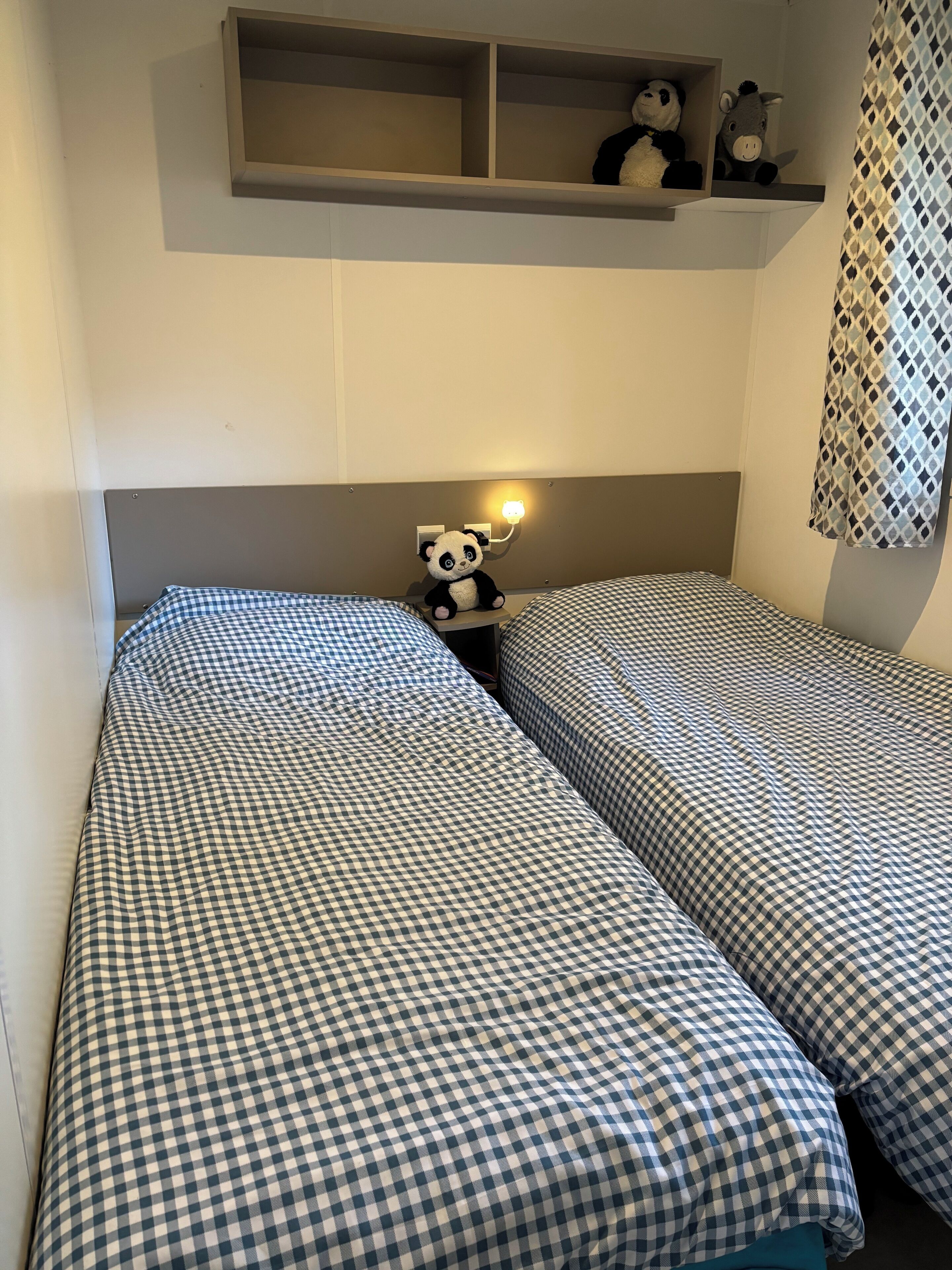 Une chambre avec deux lits simples, chacun recouvert d’une courtepointe à carreaux. Il y a une étagère au-dessus des lits, sur laquelle sont posés deux animaux en peluche, et un luminaire fixé au mur.