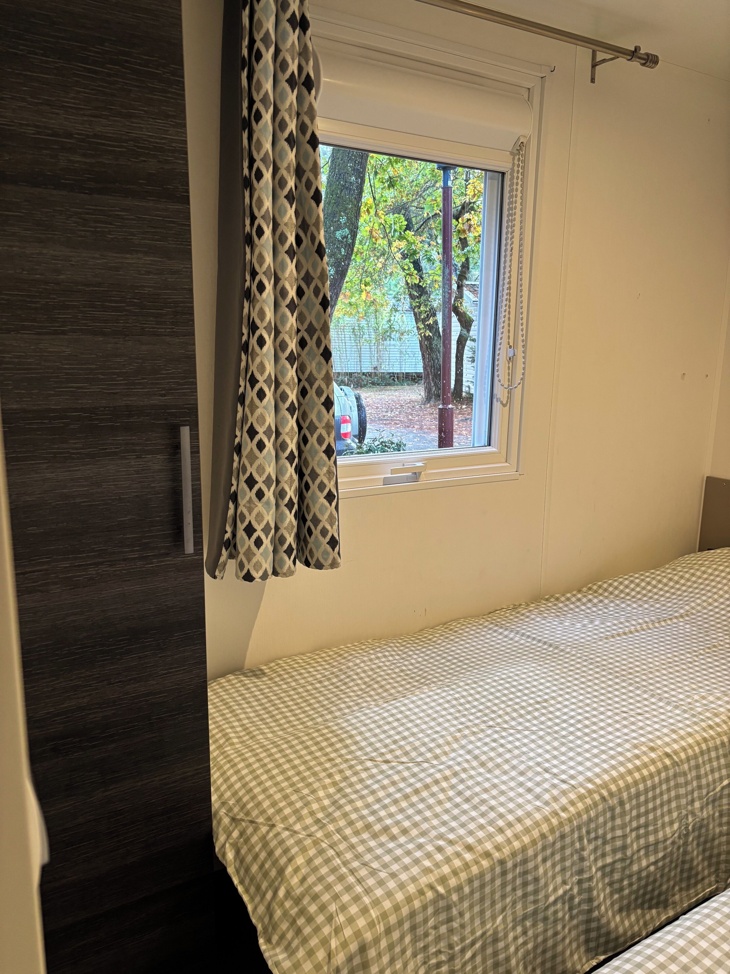 Une chambre à coucher avec une couvre-lit à carreaux, une fenêtre donnant sur des arbres et un rideau.
