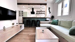 A modern living room with a flat-screen TV, a wooden coffee table, a sofa with green cushions, and a kitchen area with a bar stool and a refrigerator. - Balcony of the Sea (El Morche)
