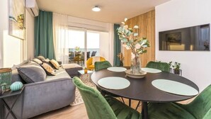 A modern living room with a sofa, green chairs, a round table, and a large window. - Blue waves apartment (Mandre)