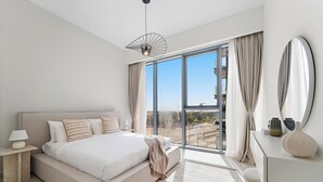 A modern bedroom with a large bed, a bedside table with a lamp, a dresser, and a view of a cityscape through floor-to-ceiling windows. - 2BR M Room CF7 B Near Jumeirah Beaches (Dubai)