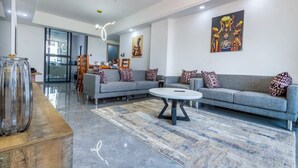 Una sala de estar moderna con un sofá gris, cojines estampados, una mesa redonda de centro y un florero de vidrio sobre una mesa de madera.