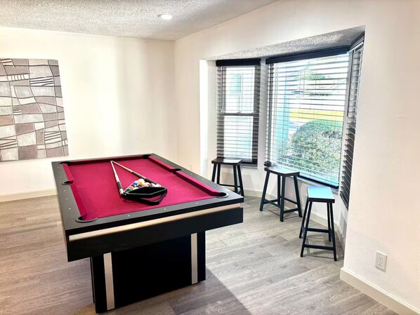 A pool table with a red felt surface, two stools, and a pool cue in a room with light wooden flooring and a large window with blinds. - Peaceful 3 Bedroom Pool Home (Tampa)