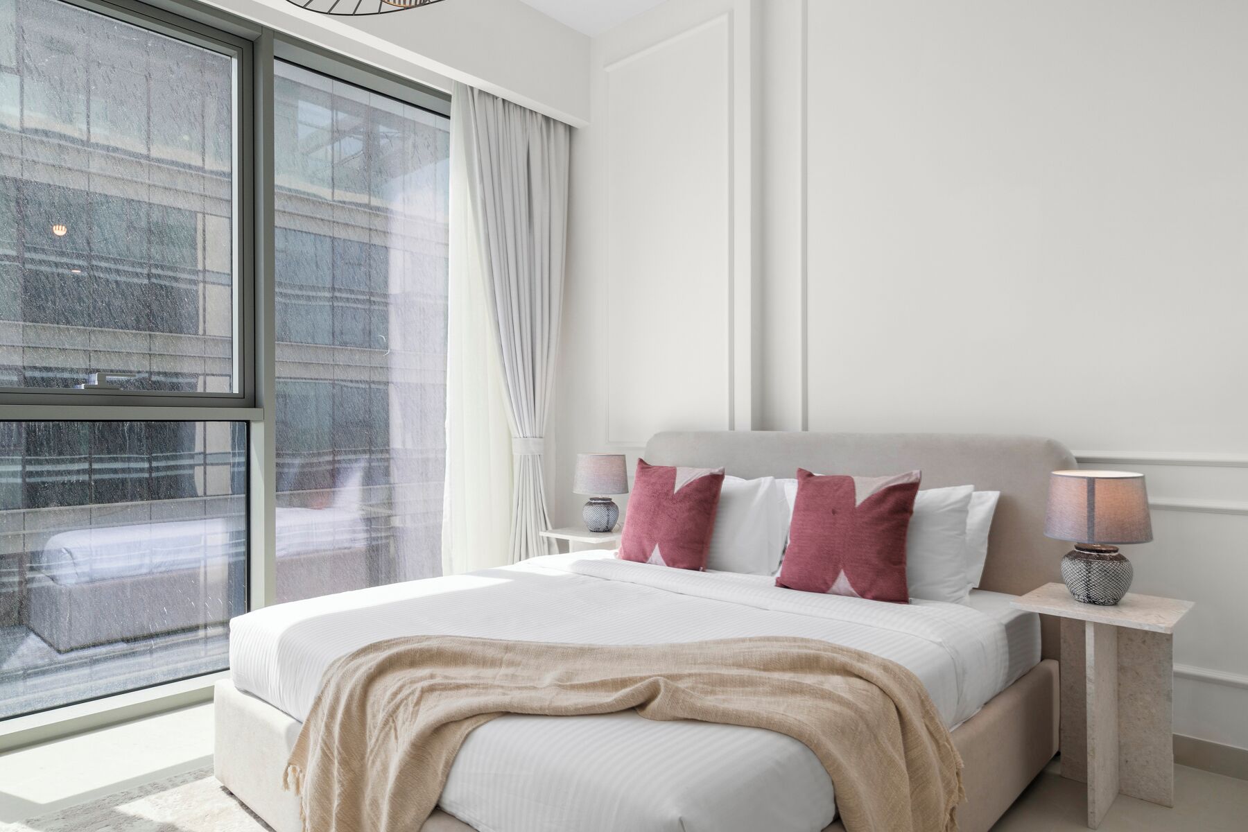 A modern bedroom with a large window, a bed with white bedding and pink pillows, and two bedside tables with lamps.