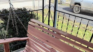 Ulkokäyttöön tarkoitettu puinen penkki, jossa on tukena ketju. Taustalla on metalliaita ja vihreää kasvillisuutta.