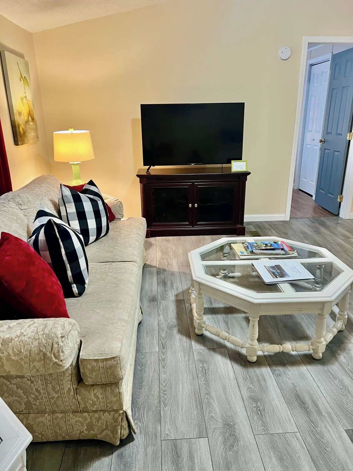 Ett vardagsrum med en beige soffa, ett soffbord i glas, ett mörkt trä TV-bord och en lampa.