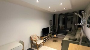 A modern living room with a flat-screen TV, a wooden chair, a sofa, a dining table, and a large window. - Cosy studio Annecy - 10 min from center and train station (Annecy)