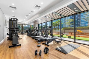 Ein modernes Fitnessstudio mit unterschiedlichen Trainingsgeräten, großen Fenstern mit Blick auf einen Park und Gebäude, sowie einem Holzboden.