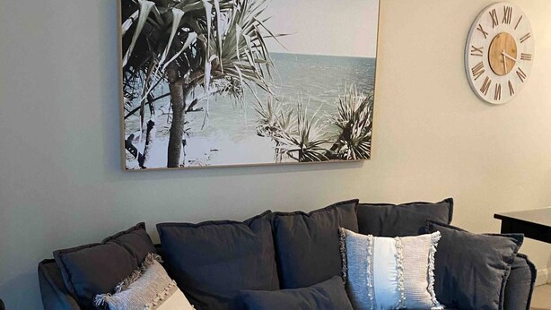 Ein dunkelbraunes Sofa mit vielen Kissen, ein gewebter Teppich, ein Holztischchen, eine Wanduhr und ein gerahmtes Bild mit einer Strandansicht.
