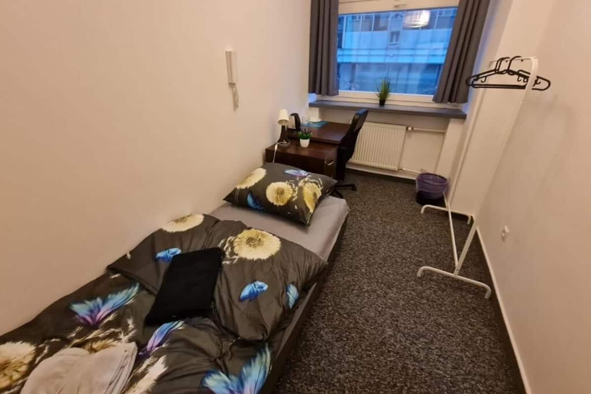 Une petite pièce, de taille modeste, comprenant un lit, un bureau et une chaise. On y trouve une fenêtre avec des rideaux, un radiateur et un porte-manteau.
