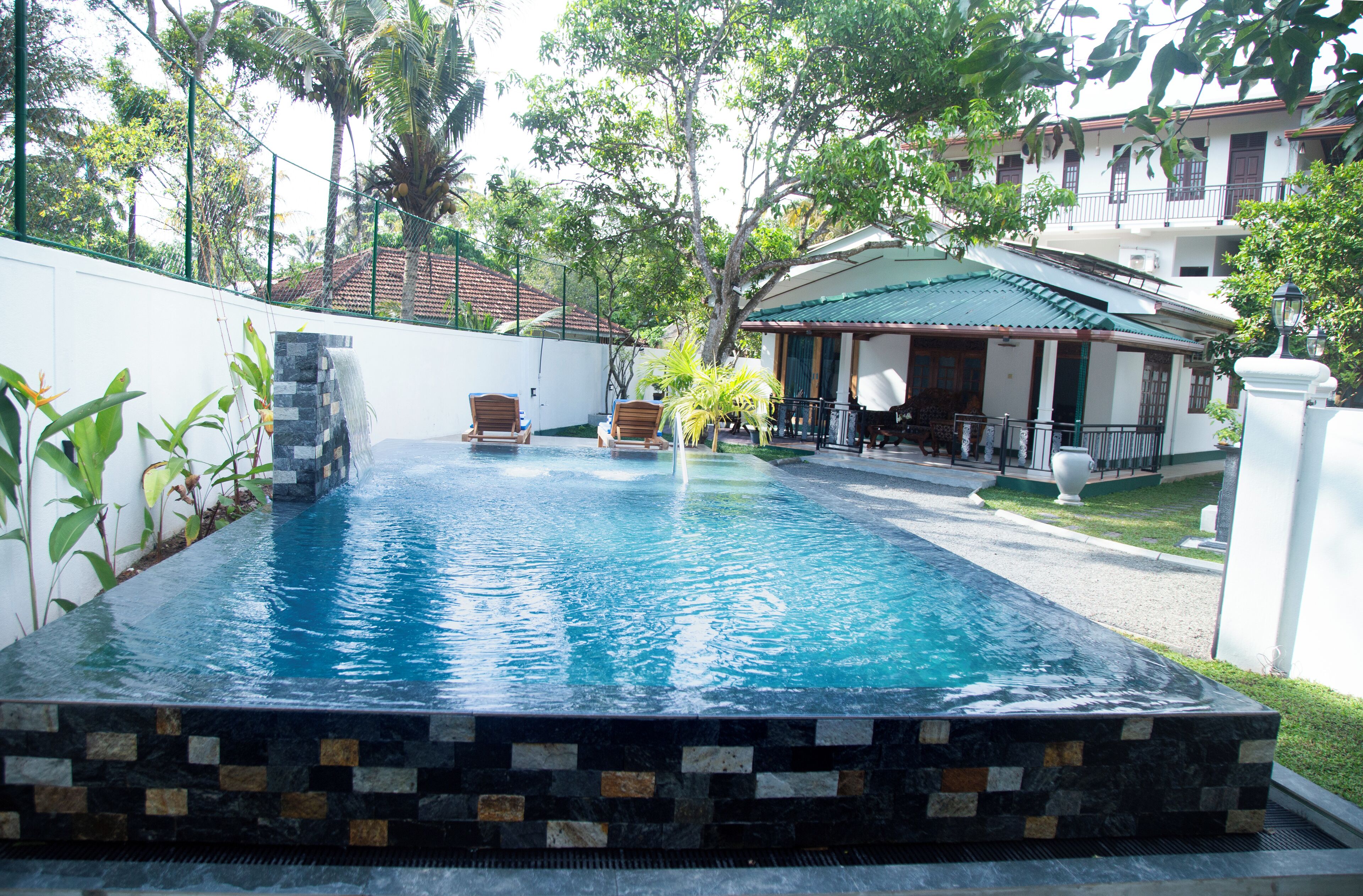 A rectangular infinity pool with a stone border, surrounded by a white wall and lush greenery.