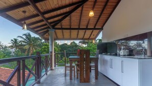 A covered outdoor dining area with a wooden table and chairs, a kitchen counter with appliances, and a view of palm trees and a red-roofed building. - Sea Bloom Villa (Thalaramba)