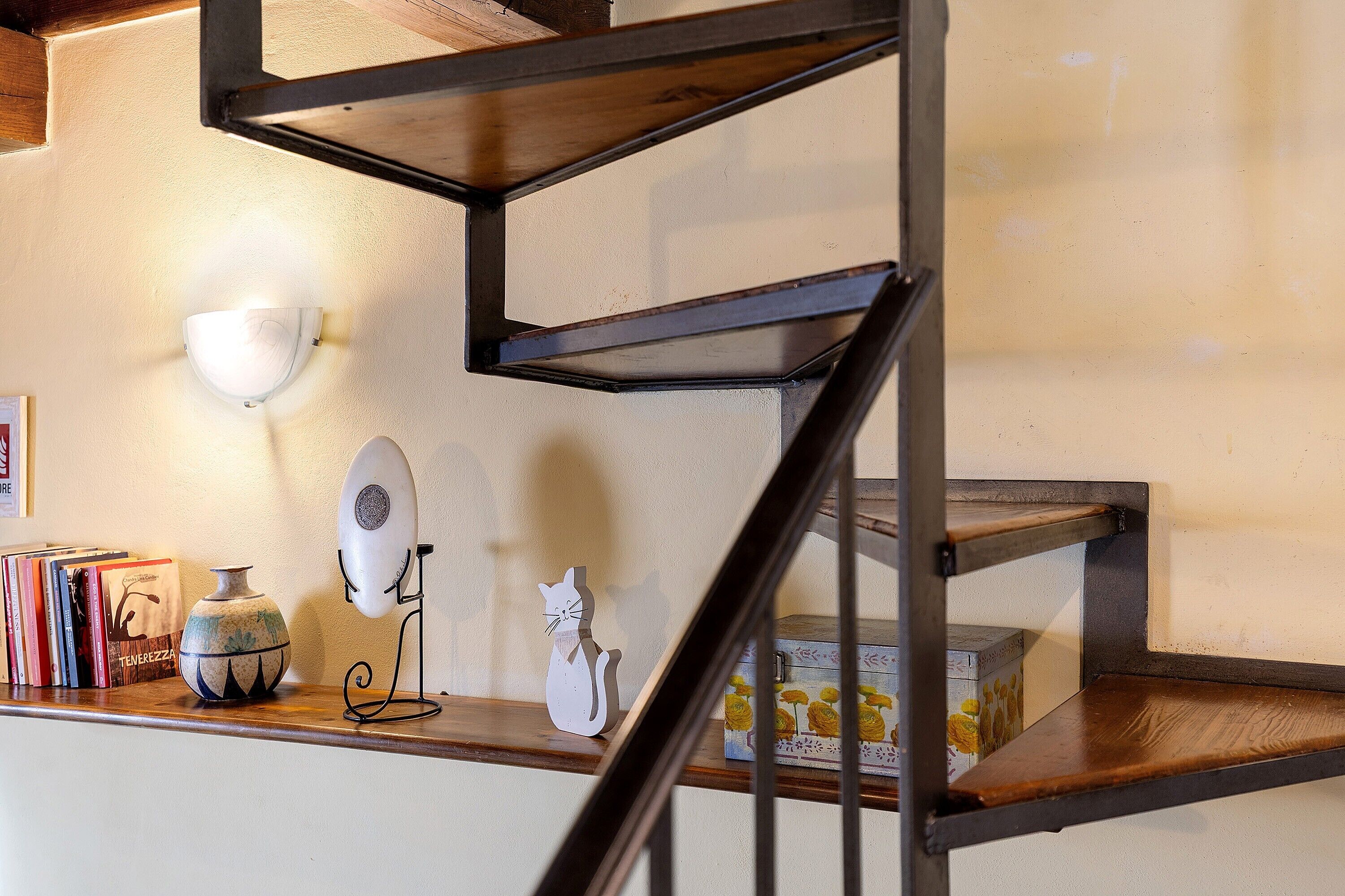 Un escalier avec une structure en métal et des marches en bois, un luminaire fixé au mur, un vase décoratif, une petite figurine de chat et une boîte de fleurs jaunes.