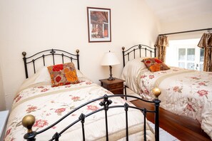 Un dormitorio con dos camas, cada una con una colcha estampada y un cojín decorativo. Hay una mesita de noche con una lámpara entre las camas, y un cuadro enmarcado en la pared.