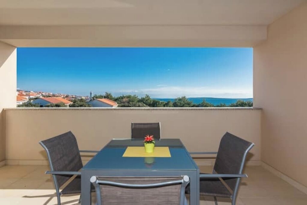 A balcony with a table and chairs, overlooking a coastal view with buildings and a clear sky.