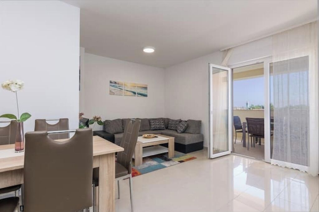 A modern living room with a sofa, a dining table, and chairs. There is a sliding glass door leading to an outdoor area.