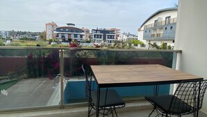Un balcón con mesa de madera y dos sillas, con vistas a una piscina y edificios modernos.
