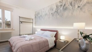 Een net opgemaakt bed met een roze dekbed, een nachtkastje met een lamp en een radiator aan de muur.