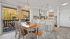 A dining area with a wooden table and white chairs, a kitchen with white cabinets and a stone backsplash, and a view of autumn foliage through a large window. - Renovated Lakefront Escape w/ Amazing Views (Garland County)