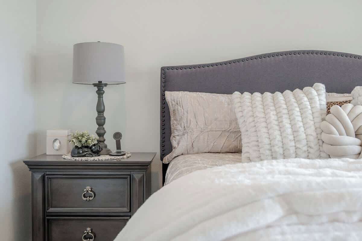 A neatly arranged bedroom with a dark wooden nightstand, a lamp, a bed with a grey upholstered headboard, and decorative pillows.