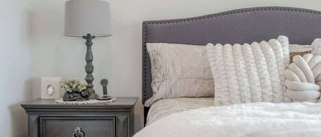 A neatly arranged bedroom with a dark wooden nightstand, a lamp, a bed with a grey upholstered headboard, and decorative pillows.