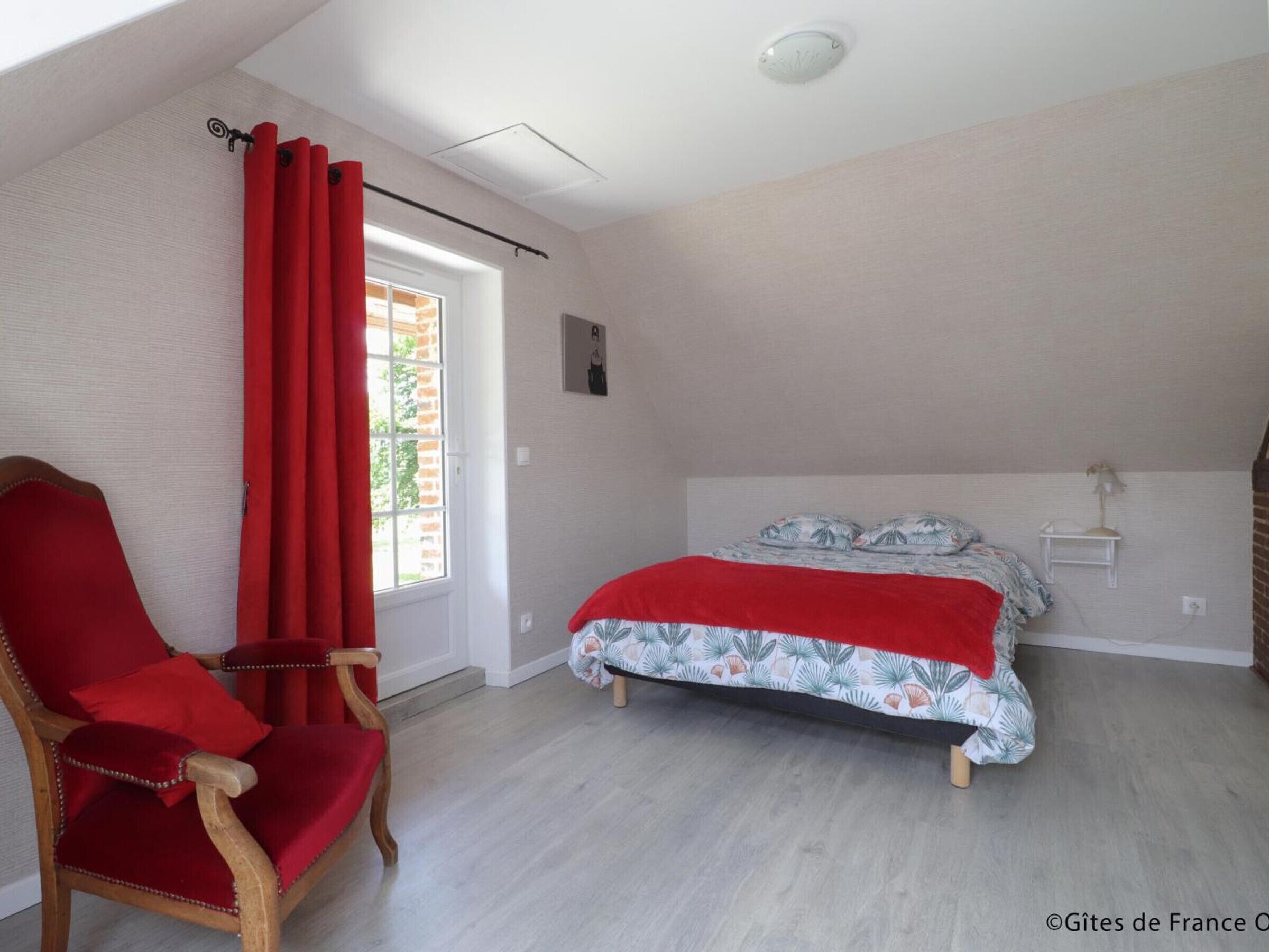 Een slaapkamer met een bed, een stoel, een raam met rode gordijnen en een klein plankje.