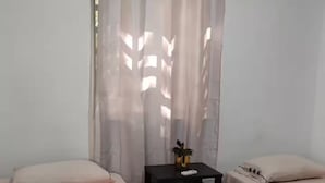 Ein Zimmer mit zwei Betten, einem kleinen, schwarzen Nachttisch und transparenten Vorhängen.