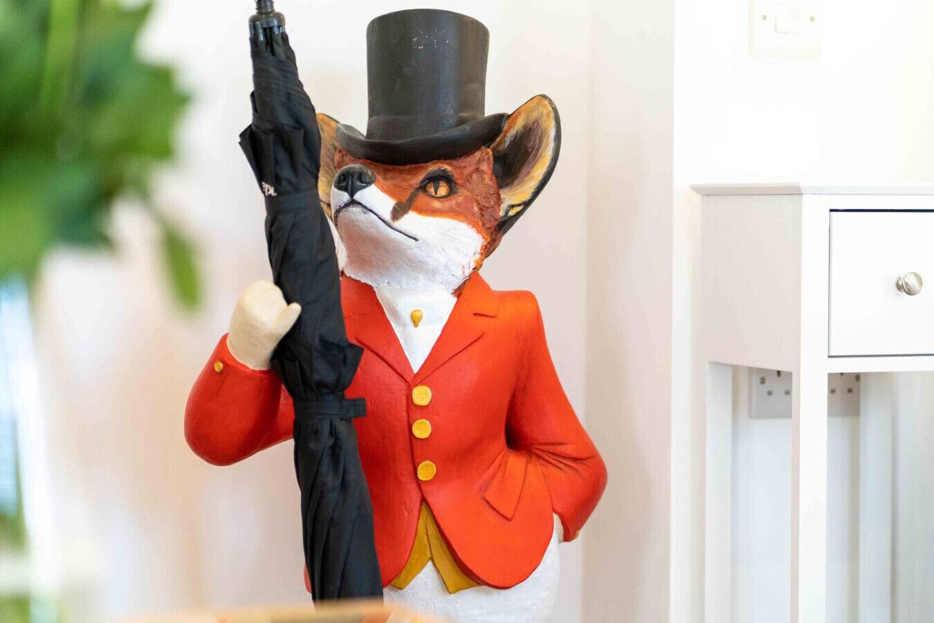 Una estatua de zorro con traje rojo y bombín negro, sosteniendo un paraguas negro.