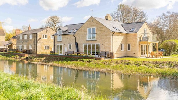 Une maison en pierre avec un étang devant, de grandes fenêtres et une terrasse aménagée avec du mobilier d’extérieur.