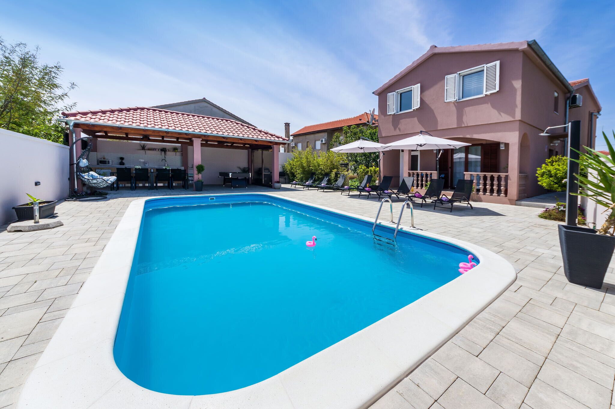 A swimming pool with lounge chairs and umbrellas, a covered outdoor kitchen, and a two-story house with a red-tiled roof.