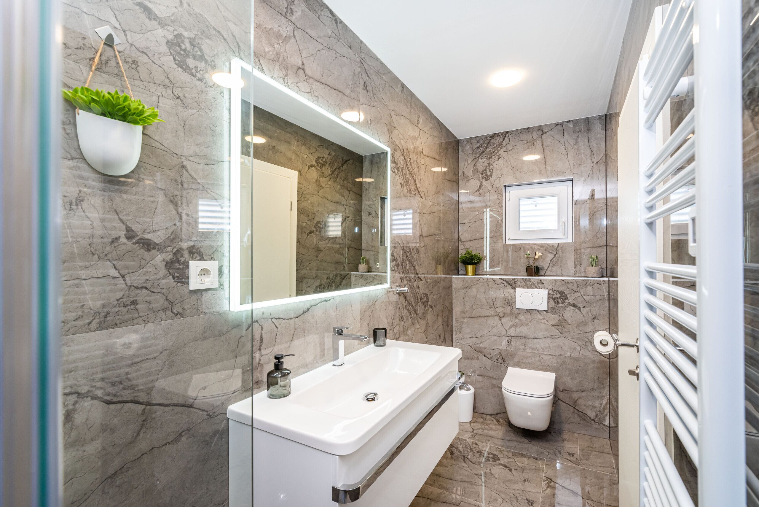 A modern bathroom with a marble wall, a large mirror, a white sink, a toilet, and a small shelf with plants.