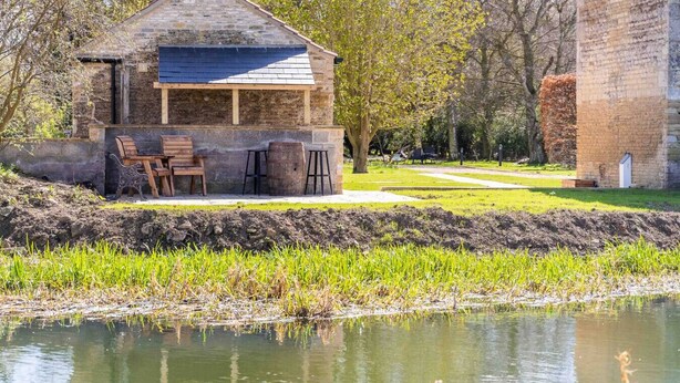 A rustic stone building with a sloped roof, wooden benches, and a small table. - The Sett: cosy riverside home (Stamford)