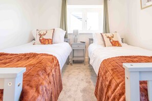 Habitación de hotel con dos camas, cada una con un cojín con motivos de ciervo, un escritorio pequeño y una ventana con cortinas.