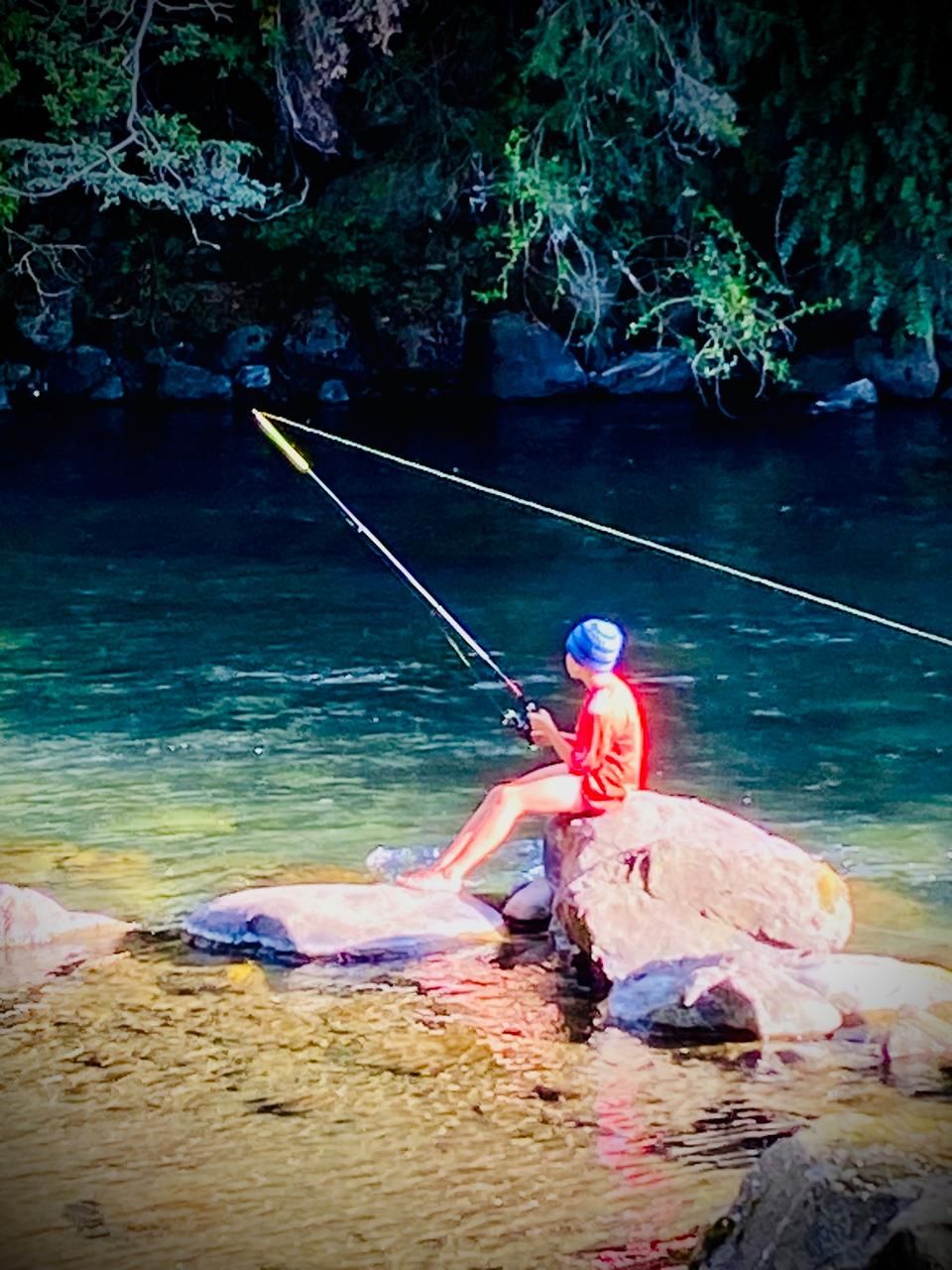 A person fishing by a clear river with rocks and greenery in the background.