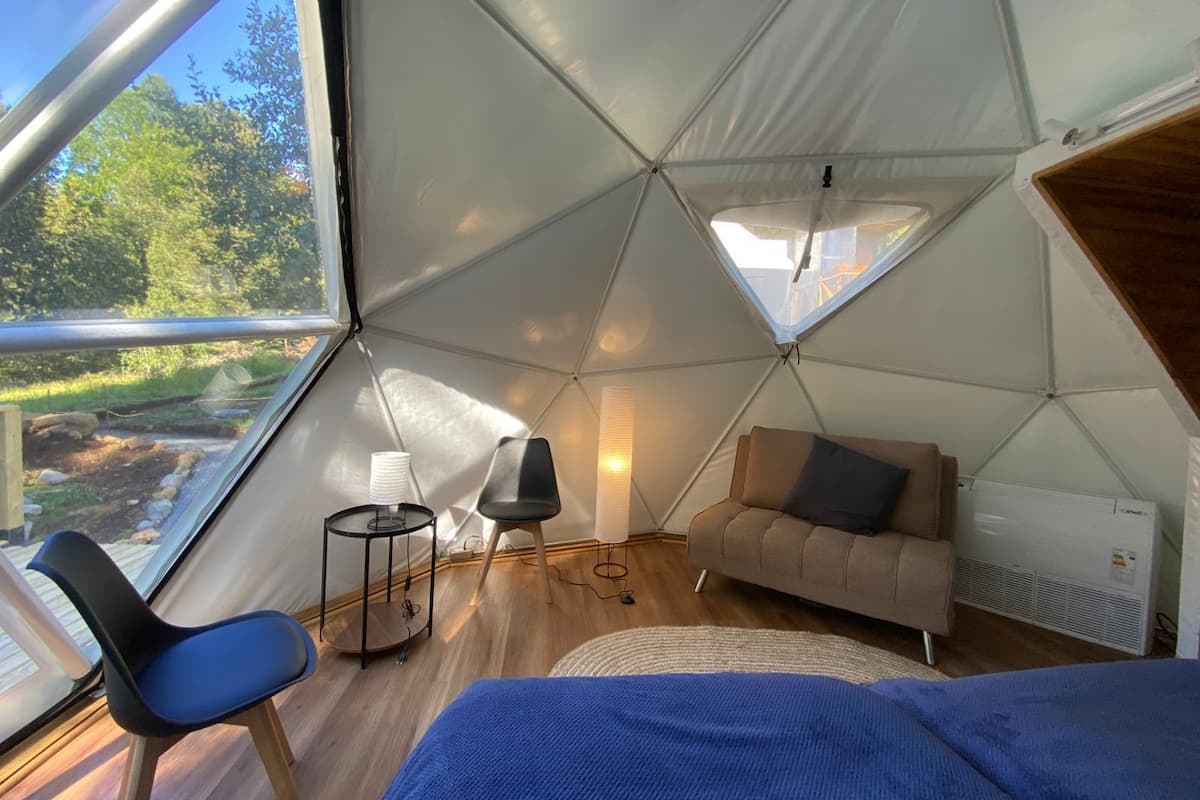 A cozy interior of a dome-shaped tent with a bed, a small sofa, a table with a lamp, and a chair.