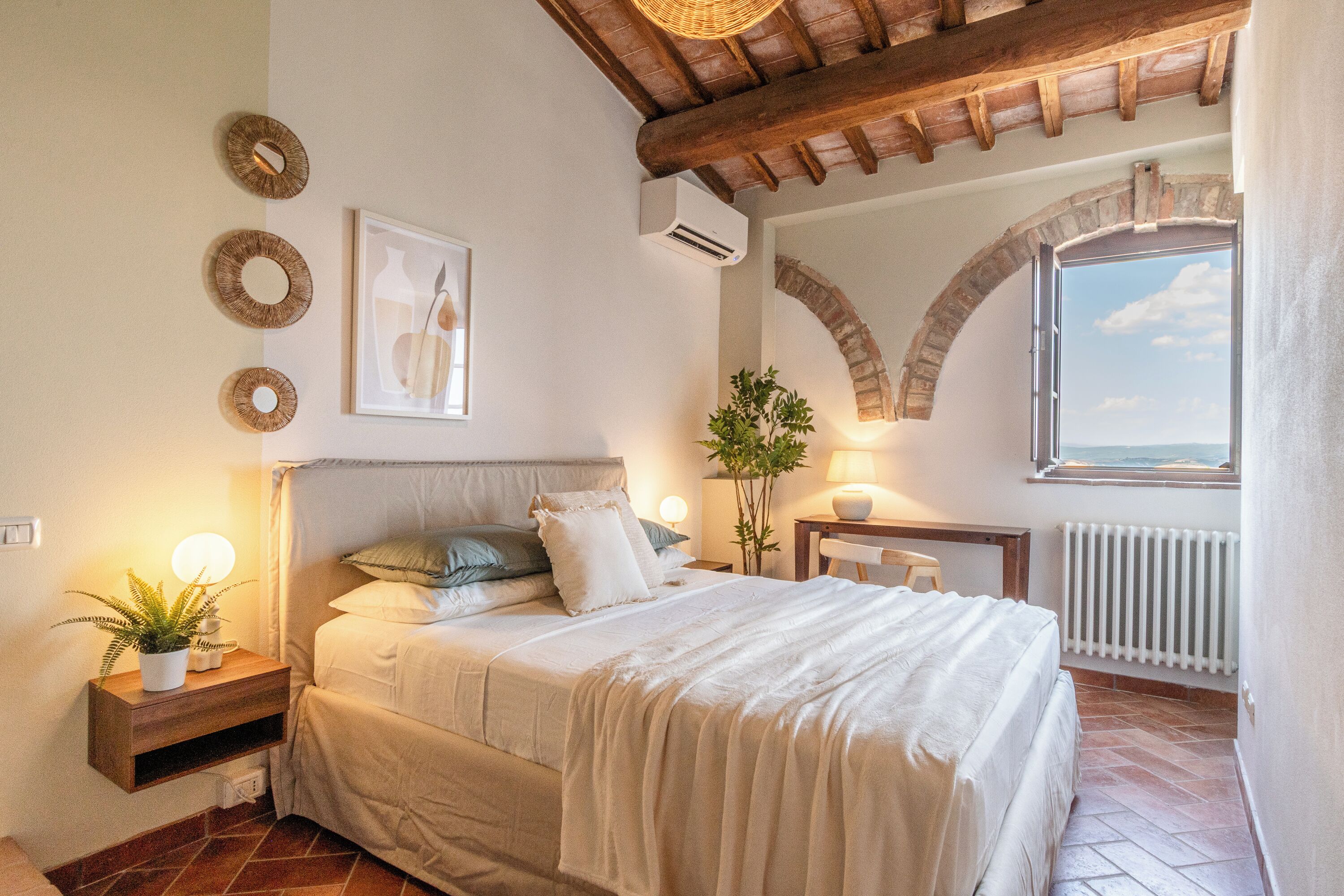 Une chambre à coucher comprenant un lit, une table de chevet, une lampe, une plante en pot et une vue sur l’extérieur par le biais d’une fenêtre en arc de cercle.