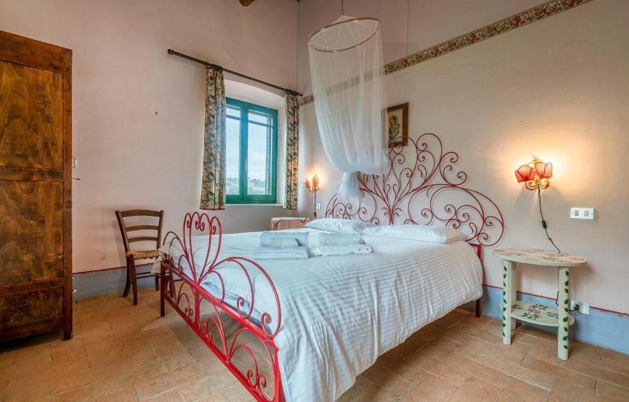 Une chambre à coucher comprenant un lit, une chaise, une table de chevet, une fenêtre avec des rideaux et un luminaire fixé au mur.