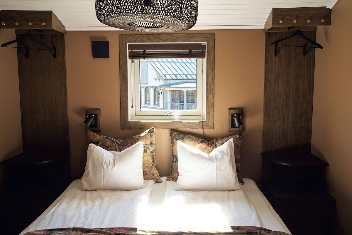 Quarto com cama, duas mesas de cabeceira, luminária pendente, janela com vista e um alto-falante fixado na parede.