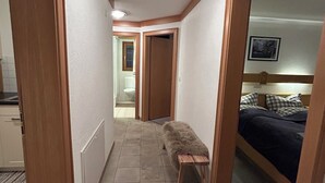 Un pasillo estrecho con un banco, puertas de madera y una cama con cabecera en la habitación contigua.