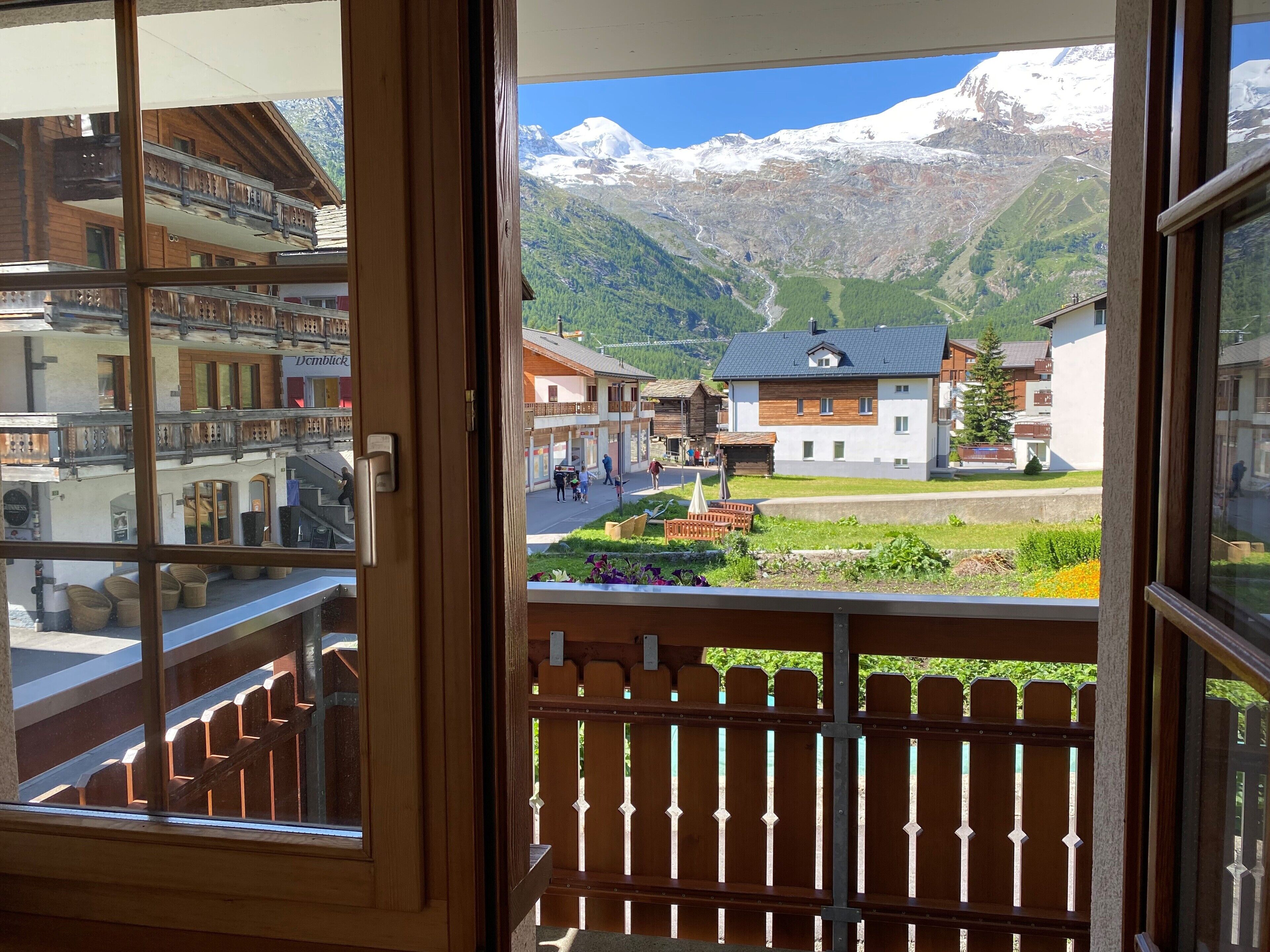 Vista da un balcone con ringhiera in legno, su una cittadina con edifici e una catena montuosa sullo sfondo.
