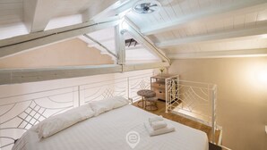 Ein Schlafzimmer mit hoher Decke, einem Bett mit weißer Bettwäsche, einem kleinen Tisch mit Stuhl und Blick auf den Himmel durch ein Dachfenster.