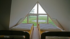 Ein Zimmer mit zwei Betten am Ende einer geneigten Decke, ein großes Fenster mit Blick auf ein grasbewachsenes Feld und ein Gewässer, sowie eine hängende Leuchte.
