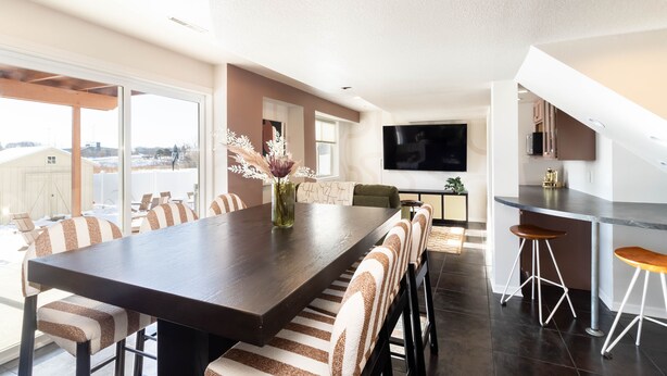A modern kitchen with a large island, bar stools, and a dining area with striped chairs. - Base Camp – Modern Cozy Retreat with Sauna, Hot Tub & Fun for Families in WDM (West Des Moines)