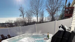 A hot tub with a glass of champagne and a bottle, surrounded by a white fence, with trees and a clear sky in the background. - Base Camp – Modern Cozy Retreat with Sauna, Hot Tub & Fun for Families in WDM (West Des Moines)