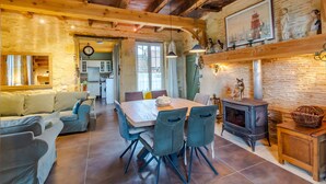 Een rustiek interieur met een eettafel, stoelen, een bank, een open haard en een keuken op de achtergrond.